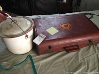 Photo showing both the enamel chamber pot with lid and the vintage brown suitcase with a round colorful sticker on top.