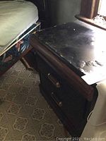 Night stand with damage on top surface seen next to a bed frame on tiled floor.