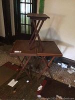 Three wooden TV trays displayed against a wall, two larger trays on the floor side by side with one smaller tray placed on top, showing foldable legs and surface condition.