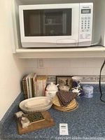 Panasonic microwave oven placed on kitchen shelf with cookbooks, dishes, trivets, kitchen linens, and cheese board on counter below