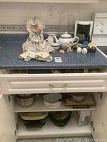 Full view of counter and open cabinet showing doll, white teapot set, coffee maker, and pots inside cabinet.