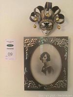 Frontal view of vintage photo of a young woman in an ornate silver metal frame beneath the decorative Venetian mask on the wall.