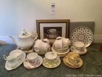 Royal Albert Val D'or teacups, saucers, various plates, cream and sugar set, and teapot with no matching lid arranged on floor with framed print and commemorative plate behind.