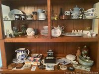 Wooden shelf units displaying a variety of decorative dishes including plates, teacups, a large pitcher, teapots, and various small ceramic decorations arranged on multiple shelves.