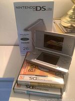 Photo of Nintendo DS Lite console, original packaging box, and two Nintendo DS games (Brain Age and Big Brain Academy) stacked together on a surface.