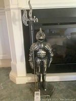 Front view of a metal knight-shaped fire utensil stand positioned in front of a fireplace, holding a pole weapon designed with detailed patterns.