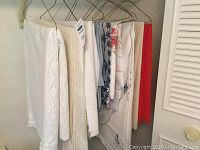 Table linens hanging on hangers showing multiple pieces of different patterns and colors including white crocheted and floral designs.