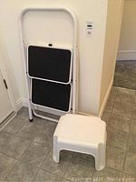 Two kitchen step ladders side by side against a wall: a white metal two-step ladder with black steps folded and a white single-step plastic stool.