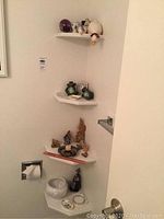 Wall corner with four white shelves displaying a mix of small ceramic and porcelain figurines including a white egg cup, two green frog figurines, and animal shapes.