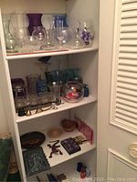 Whole shelving unit with various glass vases on top and middle shelves, assorted candle holders and brass snuffer on middle shelf, small decorative items on bottom shelf