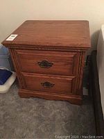 Wooden nightstand with two drawers, stained medium brown, brass decorative handles, standing on carpeted floor next to bed.