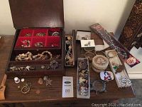 Full view of wooden jewellery box with compartments open showing assorted bracelets, rings, necklaces, watches, pill box and other costume jewellery arranged on wooden surface