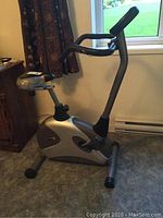 Stationary exercise bike Model 703 by Free Spirit, silver and black frame, padding on seat and backrest taped, shown in corner near window.