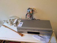 Front and angled view of the Toshiba SD-V394 combo DVD/VHS unit with power cord, remote, AV cables, and a wooden tool alongside.