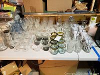 Wide view of assorted stemware and glassware on table showing clear stemmed glasses, blue tinted glasses, gold rim lowball glasses, highball glasses with newspaper text, and various other drinking glasses.