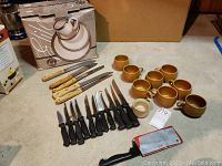 Full view of all items on a table including boxed dishes, knives with cream and black handles, stoneware mugs, and a large cleaver.