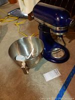 Photo showing blue KitchenAid stand mixer with stainless steel mixing bowl and three mixing attachments inside the bowl on a concrete floor.
