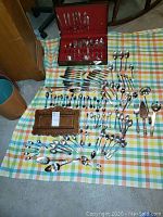 Wide view of the entire assortment of silverplate utensils including the boxed spoons, loose spoons, forks, knives, and the wooden hanging spoon rack arranged on a checkered cloth.