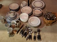 Full collection of dishes and silverware arranged on concrete floor, showing stacks of plates, bowls, mugs, cups, and a basket of silverware.