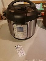 Full view of the Instant Pot Duo Plus electric pressure cooker showing stainless steel body and black lid with handle, placed on a table with a MaxSold tag nearby.