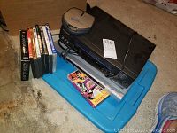 Photo showing VCR player, DVD player, Sony Discman CD player and DVDs arranged on a blue plastic container lid on a concrete floor