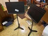 Two black Manhasset metal music stands standing on beige floor in a room with books and other items around.
