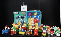 Photo showing M&M's lunch pail, Happy Lights card with 4 purple M&M's figures, and 10+ small M&M's character figures on toy vehicles and floats arranged in a semi-circle.