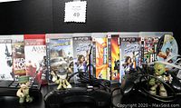 Photo showing 12 PS3 game cases lined up, three small action figures holding weapons, seven black controllers, and assorted cables and connectors on a black surface.
