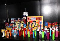 Front view of assembled vintage PEZ dispensers with colorful character heads, candy packs and PEZ Power magazine arranged behind on black shelf.