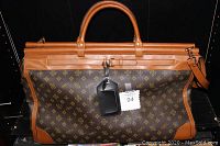 Front view of the overnight bag showing brown leather trim, carry handles, lockable zipper top, footed bottom, and printed dark brown body with design pattern.