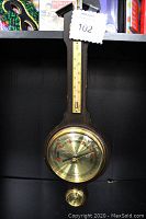Front view of weather instrument showing wood base with brass thermometer at top, large brass barometer in center, and brass relative hygrometer at bottom. Includes a tag with lot number.