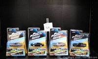 Four unopened Jada Fast & Furious diecast cars from 2016, two grey Chevy Chevelle SS, one black Dodge Charger R/T, and one black Dodge Challenger SRT8 with official movie branding and extra parts to build a Mitsubishi Lancer Evolution VII included in packaging.