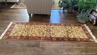 Long beige area rug runner with red border and floral motifs on wood floor, visible fringe on short edges