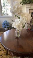 Full view of the silk flower arrangement with large white blooms in a clear glass vase placed on a wooden table.