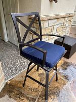 Black metal outdoor bar stool with navy blue cushion shown from side angle, showing armrest and swivel base on stone patio.