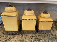 Three yellow ceramic square canisters with ornate bronze bases and lids arranged on countertop.