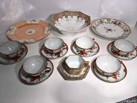 Overview of six Geisha Girl teacups with saucers, Nippon moriage bowl and plate, and Royal Rochester plate arranged on white backdrop.