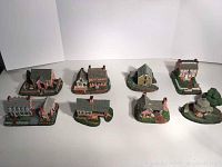 Wide shot of all eight miniature church sculptures arranged in two rows on white surface, showing detailed colonial architecture and greenery.