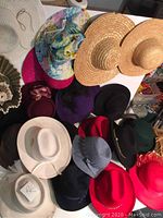 Top-down view of 15 various women's hats displayed on a white table, showing different styles and colors including straw, felt, and fabric hats.