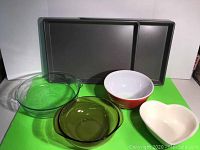 Photo showing two rectangular baking sheets laid horizontally, four bowls placed in front on green surface: clear large Pyrex, amber medium Pyrex, white heart-shaped Pfaltzgraff, and orange-white Fire King bowl.