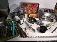 Kitchen appliances and utensils on table including toaster, blender, knife sharpener, metal and plastic utensils