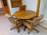 Photo showing octagonal oak kitchen table with the leaf included, two rolling chairs surrounding it.