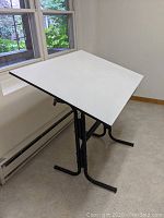 Front angle view of the drafting table showing the white laminate top and black steel legs positioned near a window.