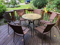 Full view of round patio table with 6 matching brown woven stacking chairs arranged around it on wood deck.