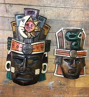 Pair of two Mexican wood masks on a wooden surface, showing the larger mask with sun and moon motif and the smaller mask with green serpent design.
