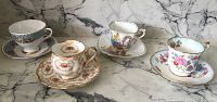 Four vintage tea cups and saucers displayed on a marble surface, showing different floral patterns and gold accents.