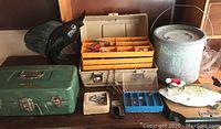 All fishing-related items arranged including green tackle box, beige tackle box with fishing lures, metal minnow pail, various decorative fish figures, and small plastic boxes.