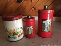 Front view of three vintage metal canisters: one white floral canister and two red labeled 'T' and 'F' canisters