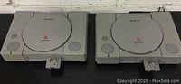 Two Sony PlayStation 1 consoles side by side, both with game cartridges inserted and showing PlayStation logos.