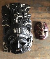 Photo showing two wooden masks on a wooden surface: one large black and white carved mask (16 inches) with a tribal design, and a smaller red mask with a turtle motif on the forehead